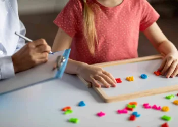 Unrecognizable little girl exercising with alphabet board during session with female child development specialist at clinic, small kid making words from colorful plastic letters, cropped image