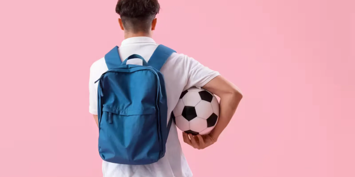 Male student with backpack and soccer ball on pink background, back view