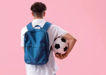 Male student with backpack and soccer ball on pink background, back view