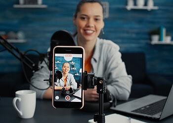 Attractive social media influencer sitting at home studio desk while starting streaming from living room with smartphone. Young content creator recording daily vlog with touchscreen modern device.